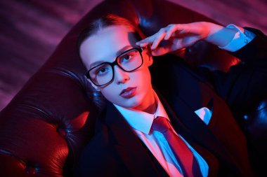 Deri koltukta kravat ve gözlük takmış şık, kendine güvenen genç bir kadının fotoğrafı. Karışık ışıklandırma. Lüks bir iş tarzı. Gözlük modası.