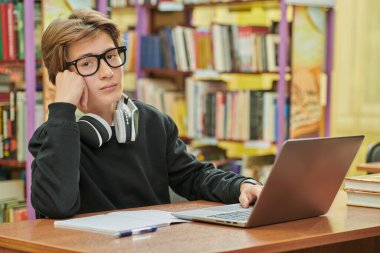 Gözlüklü ve kulaklıklı zeki bir öğrenci kütüphanede bir masada oturup dizüstü bilgisayarla çalışırken kameraya bakar. Eğitim. Modern öğrenciler.