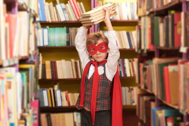 Okul üniforması giymiş bir çocuğun portresi ve kitaplıkların arasında duran süper kahraman kırmızı pelerin ve maske. Eğitimde süper yetenekler. Çocukların rüyaları. 