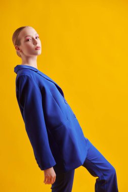 Moda ve güzellik. Yumuşak pembe makyajlı güzel bir manken sarı stüdyo arka planında zarif mavi bir takım elbiseyle poz veriyor. Parlak yaz renkleri. 