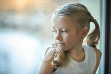 Beyaz tişörtlü şirin bir kızın portresi pencere pervazında oturuyor ve pencereden gülümsüyor. Karlı kış günü. Çocuklar, çocukluk ve duygular. Boşluğu kopyala.