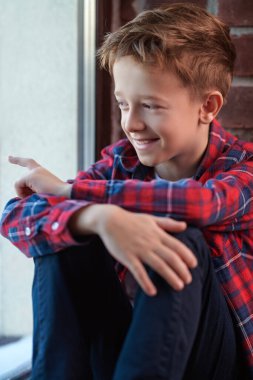 Ekose gömlekli ve kotlu mutlu bir sarışın çocuk pencere eşiğinde tuğladan bir duvara yaslanıyor. Çocuklar ve çocukluk. 