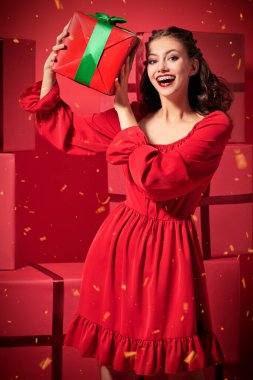 Güzel kırmızı elbiseli, akşam makyajı ve saç stili olan güzel bir kız neşe içinde bir hediye kutusu tutuyor. Bir sürü kırmızı hediye kutusu var. Noel ve Yeni Yıl hediyeleri. 