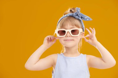 Çocuklar, mutlu çocukluklar. Yazlık kıyafetleri ve şık güneş gözlükleriyle beş yaşında şirin bir kız kameraya bakıp gülümsüyor. Çocuk modası. Metin için alanı olan sarı stüdyo arkaplanı.