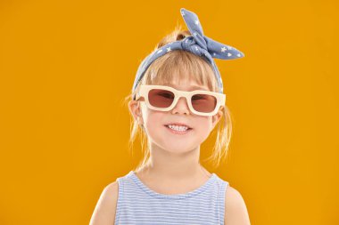 Çocuklar, mutlu çocukluklar. Yazlık kıyafetleri ve şık güneş gözlükleriyle beş yaşında şirin bir kız kameraya bakıp gülüyor. Çocuk modası. Metin için alanı olan sarı stüdyo arkaplanı.