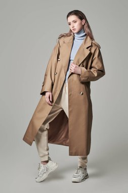 Gri bir arka planda bej renkli trençkot giyen çekici, kendine güvenen bir mankenin tam boy fotoğrafı. Kadın modası. Sonbahar-ilkbahar.