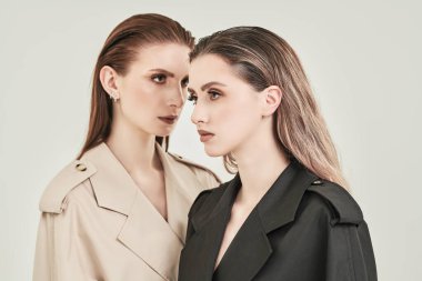 Beyaz bir stüdyo arka planında moda trençkotlarla poz veren iki şık güzel kız mankeni. Kadın modası ve güzelliği.