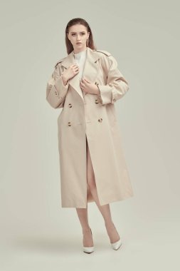 Şık yarı sezon stili. Beyaz trençkotlu ve yüksek topuklu deri ayakkabılı sevimli genç bir kadının tam boy portresi. Beyaz stüdyo geçmişi. Kadın modası.