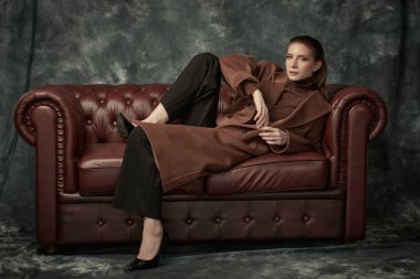 Çekici, kendine güvenen bir kadın deri koltukta poz veriyor moda keçeli ceketi, siyah pantolonu ve yüksek topuklu ayakkabıları var. Kadın modası.