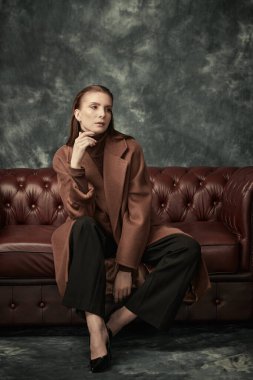 Çekici, kendine güvenen bir kadın deri koltukta poz veriyor moda keçeli ceketi, siyah pantolonu ve yüksek topuklu ayakkabıları var. Kadın modası.