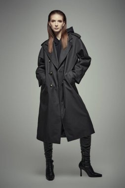 Siyahlı kadın. Siyah trençkotlu ve yüksek topuklu deri çizmeli şık, kendine güvenen bir kadının tam boy portresi. Gri stüdyo geçmişi. Kadın modası.