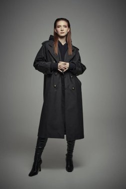 Siyah trençkotlu ve yüksek topuklu deri çizmeli şık, kendine güvenen bir kadının tam boy portresi. Gri stüdyo geçmişi. Kadın modası.