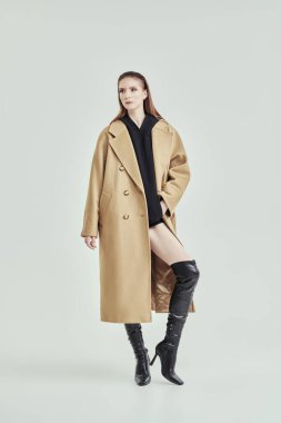 Bej renkli trençkotlu ve yüksek topuklu siyah çizmeli şık, kendine güvenen bir kadının tam boy portresi. Beyaz stüdyo geçmişi. Kadın modası.