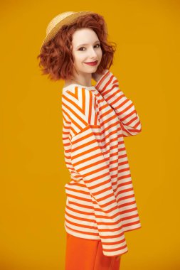 Kıvırcık saçlı, saman şapkalı ve turuncu çizgili uzun kollu sarı stüdyo arka planında poz veren neşeli, güzel bir kız. Olumlu duygular. Yaz modası ve güzelliği.