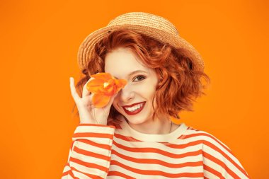 Kıvırcık saçlı neşeli bir kız kameraya geniş bir şekilde gülümsüyor ve bir gözünü orkide çiçeğiyle kaplıyor. Turuncu stüdyo geçmişi. Mutlu hayat resimleri. Yaz havası. 