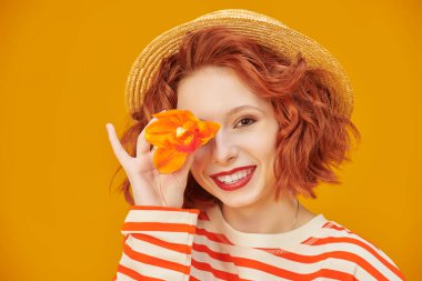 Yaz havası. Kıvırcık saçlı neşeli bir kız kameraya geniş bir şekilde gülümsüyor ve bir gözünü orkide çiçeğiyle kaplıyor. Fotokopi alanı olan sarı stüdyo. Mutlu hayat resimleri.