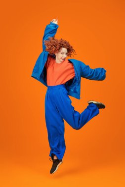 Mavi ve turuncu giysiler içinde atlayan parlak kırmızı kıvırcık saçlı, neşeli model kızın tam boy portresi. Parlak renkler. Stüdyo turuncu arka plan. Yaz modası. 