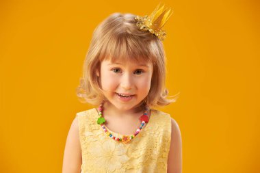 Sarı saçlı, sarı elbiseli ve altın tacı olan, neşeyle gülen şirin küçük bir prensesin portresi. Sarı stüdyo geçmişi. Mutlu çocukluk, mutlu çocuk duyguları.