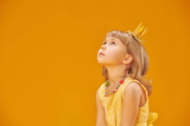 Sarı elbiseli ve altın tacı takmış güzel küçük bir prenses rüya gibi yukarı bakıyor. Mutlu bir çocukluk. Çocukların fantezileri ve hayalleri. Peri masalları. Sarı arka planda stüdyo portresi. Boşluğu kopyala.