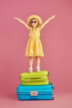 Mutlu yaz tatilleri! Parlak sarı elbiseli ve hasır şapkalı şirin bir kız parlak bavulların üzerinde mutlu bir şekilde duruyor. Pembe arka planda stüdyo portresi. Metin için yer.