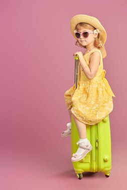 Tatile çık! Parlak sarı bir yaz elbisesi ve hasır şapkalı şirin bir kız yeşil bir bavulla mutlu bir şekilde poz veriyor. Pembe arka planda stüdyo portresi. Metin için yer.