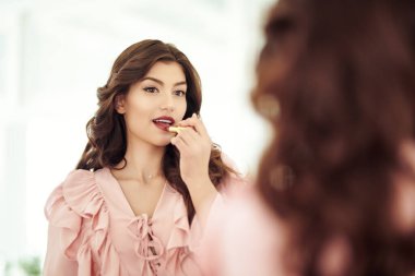 Uzun kıvırcık saçlı ve çekici gülüşlü güzel bir kadın güneşli yatak odasında aynanın önünde makyaj yapıyor. Mutlu hayat, mutlu insanlar konsepti. Makyaj. 