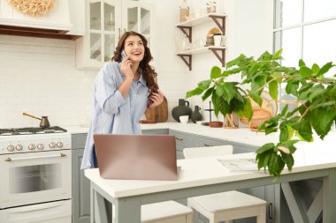 Evinin mutfağında güzel, mutlu bir genç kadın akıllı bir telefonla konuşuyor ve gülümsüyor. Mutlu bir hayat. Konforlu ev kavramı. 