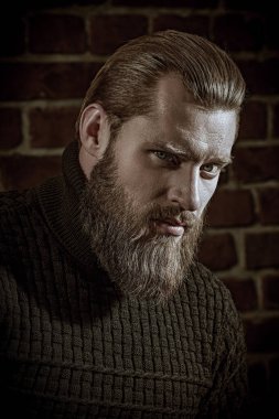 Cesaret ve güç. Büyük sakallı bir adamın portresi, örgü örülmüş bir süveter içinde, tuğla bir duvarın arkasında duran ve sakince kameraya bakıyor..