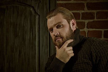 Cesaret ve güç. Büyük sakallı bir adamın portresi, örgü örülmüş bir süveter içinde, tuğla bir duvarın arkasında duran ve sakince kameraya bakıyor..