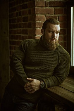 Örgü kazaklı yakışıklı bir adamın portresi, tuğla bir duvarın arkasında duruyor ve sakince ve düşünceli bir şekilde pencereye bakıyor..