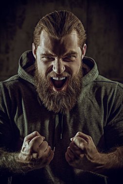 Siyah kazaklı, yumruklarını sıkan ve öfkeyle bağıran iri yarı, sakallı bir adamı yakından çek. Erkeklik ve fiziksel güç kavramı..