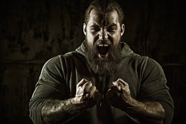 Siyah tişörtlü, yumruklarını sıkan ve öfkeyle bağıran iri yarı, sakallı bir adam. Erkeklik ve fiziksel güç kavramı..