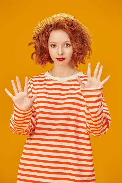 Kıvırcık kızıl saçlı güzel bir kız, hasır şapka giymiş ve sarı stüdyo arka planında turuncu çizgileri olan uzun kollu bir kız. İnsanların duyguları. Yaz modası ve güzelliği.