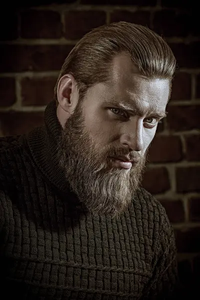 Cesaret ve güç. Büyük sakallı bir adamın portresi, örgü örülmüş bir süveter içinde, tuğla bir duvarın arkasında duran ve sakince kameraya bakıyor..