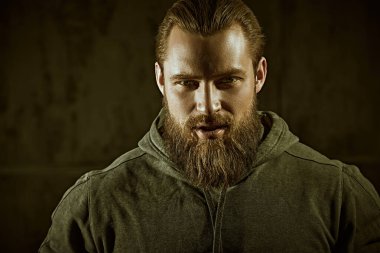 Siyah kapüşonlu, beton duvara dayanmış iri ve vahşi sakallı bir adamın yakın plan portresi. Erkeklik ve fiziksel güç kavramı..