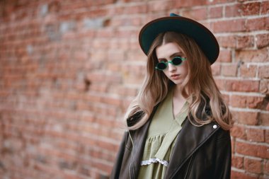 Şık güneş gözlüklü, şapkalı ve deri ceketli güzel hippi kız şehrin duvarında duruyor. Boho ve hipster tarzı. İnsanlar ve yaşam tarzı. Boşluğu kopyala.