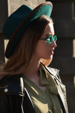 Şık güneş gözlüklü, şapkalı ve deri ceketli güzel hippi kız güneşe bakan bir caddede duruyor. Boho ve hipster tarzı. İnsanlar ve yaşam tarzı.