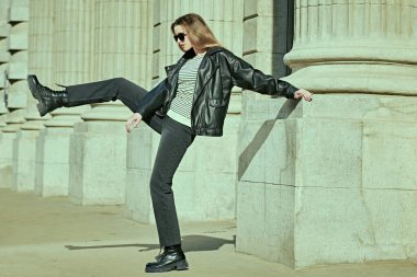 Uzun menzilli çekim. Siyah deri ceketli, siyah kot pantolonlu ve güneş gözlüklü modern bir kadın. Deri kıyafet modası. Çekici bir genç kadın. Moda ve gençlik tarzı. 