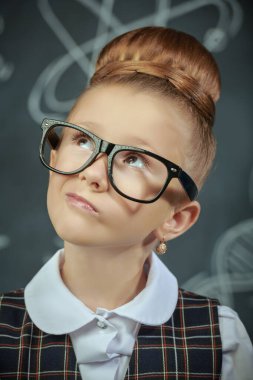 Eğitici bir kavram. Okul üniformalı ve gözlüklü şirin bir kızın portresi şüpheli bir şekilde yukarı bakıyor. Bilimsel çizimleri olan siyah bir kara tahta. Genç bilim adamları.