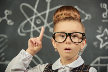 Okul üniformalı ve gözlüklü sevimli zeki bir kız dikkat çekmek için işaret parmağını kaldırıyor ve kameraya bir sürprizle bakıyor. Bilimsel çizimleri olan siyah bir karatahta. Eğitici kavram. 