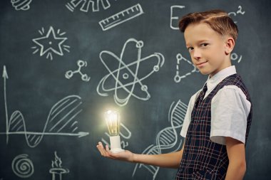 Çocukların erken gelişimi. Okul kıyafetli zeki bir çocuk elinde yanan bir ampul tutuyor ve gülümsüyor. Tebeşirle çizilmiş siyah okul panosunun arka planı. Keşifler. Eğitim.