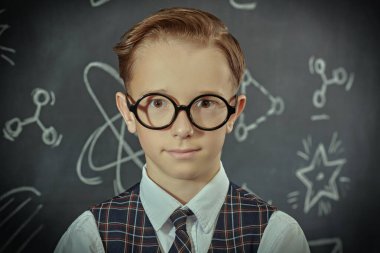 Okul üniformalı, gözlüklü ve kameraya ciddi ciddi bakıp gülümseyen zeki bir çocuğun portresi. Bilimsel çizimleri olan siyah bir kara tahta. Eğitim. Genç bilim adamları.