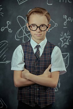 Okul üniformalı, gözlüklü ve kameraya ciddi ciddi bakıp gülümseyen zeki bir çocuğun portresi. Bilimsel çizimleri olan siyah bir kara tahta. Eğitim. Genç bilim adamları.