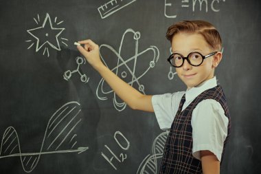 Okul üniformalı, gözlüklü ve kameraya ciddi ciddi bakıp gülümseyen zeki bir çocuğun portresi. Bilimsel çizimleri olan siyah bir kara tahta. Eğitim. Genç bilim adamları.