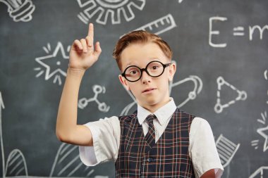 Okul üniformalı, gözlüklü ve kameraya ciddi ciddi bakan zeki bir çocuğun portresi. Bilimsel çizimleri olan siyah bir kara tahta. Eğitim. Genç bilim adamları.