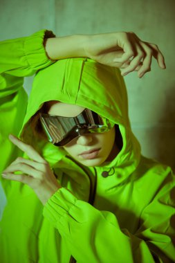 Modern hip-hop dansçısının portresi. Yeşil kapüşonlu ve güneş gözlüklü şık bir kız stüdyoda poz veriyor. Gençlik tarzı.