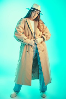 Moda, yarı sezon kıyafetleri ve aksesuarlar. Güzel esmer bir kızın tam boy portresi şık kahverengi bir trençkot, geniş bir kot pantolon ve mavi bir arka planda beyaz bir bluz giyiyor.. 