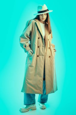 Moda, yarı sezon kıyafetleri ve aksesuarlar. Güzel esmer bir kızın tam boy portresi şık kahverengi bir trençkot, geniş bir kot pantolon ve mavi bir arka planda beyaz bir bluz giyiyor.. 