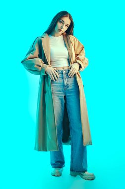 Moda, yarı sezon kıyafetleri ve aksesuarlar. Güzel esmer bir kızın tam boy portresi şık kahverengi bir trençkot, geniş bir kot pantolon ve mavi bir arka planda beyaz bir bluz giyiyor.. 