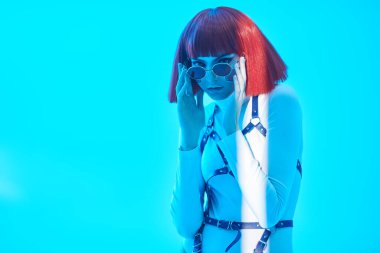 Sanal oyunlar. Siber punk kavramı. Parlak kırmızı makyajlı, kırmızı saçlı güzel bir siber punk kızın portresi. Mavi stüdyo arka planına karşı beyaz tulum içinde poz veriyor. Makyaj ve saç stili. 